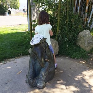 small girl sitting on small statue of elephant, with a hat on its head