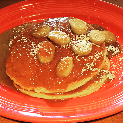 plate of pancakes with caramel sauce, bananas, and powdered sugar