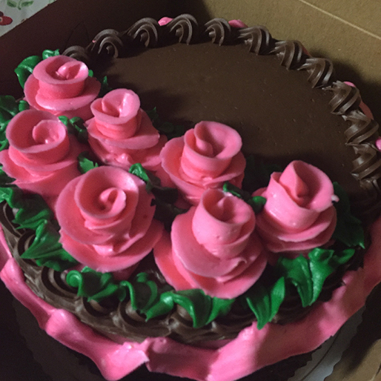Cake covered in chocolate frosting with pink frosting roses and green frosting leaves, plus a ribbon of pink frosting around the upper edge