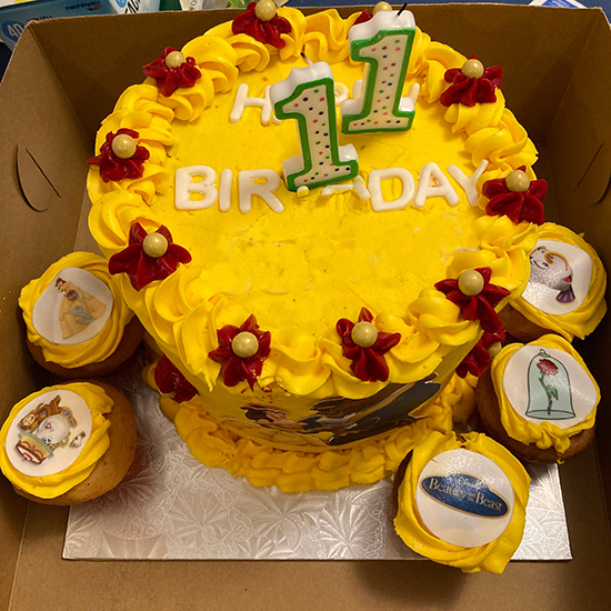 elaborate yellow cake with red frosting flowers and two #1 candles surrounded by cupcakes topped with images from Beauty and the Beast in fondant