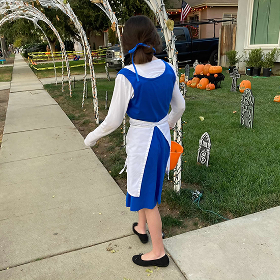 girl with brown hair tied with blue ribbon trick-or-treating while wearing white shirt, sleeveless blue dress, white apron, and black shoes