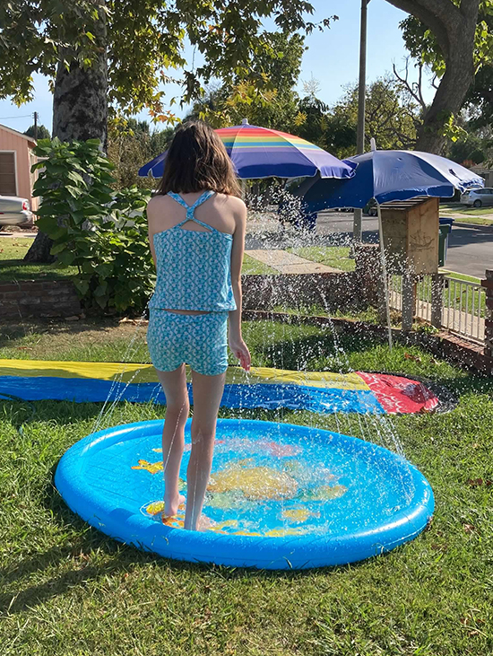 girl with brown hair in blue and white tankini standing in splash pad on lawn