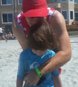 Man in running gear hugging a little girl in a blue rashguard top; both are on the beach with a building in the background