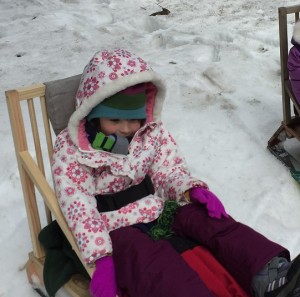 Little girl in flowered winter coat, hat, and mittens sitting on a wooden sled with a high back and seat belt