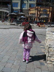 Little girl in flowered coat and large purple-and-black striped scarf in a stone patio with chairs and umbrellas in the background